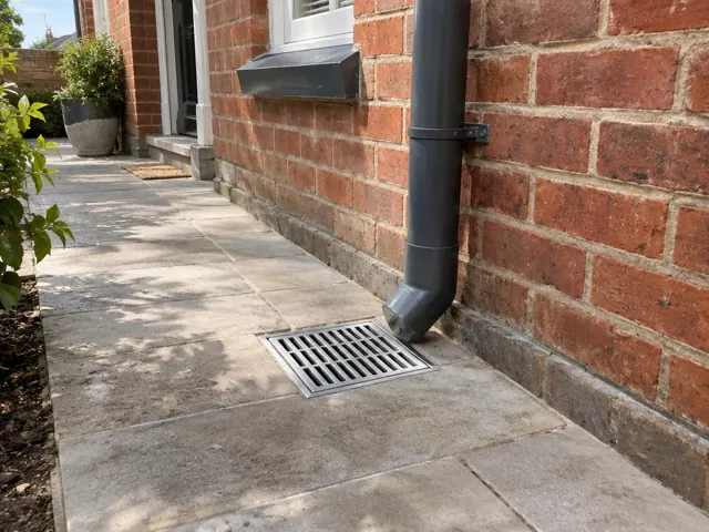 Outdoor gully grate beside a brick house wall and downpipe on a dry paved path in Clovelly